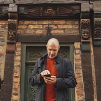 A man is standing in front of a half-timbred house. He is looking at his phone