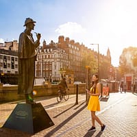 A young woman in a yellow dress is standing in front of the Sherlock Holmes statue on Baker Street