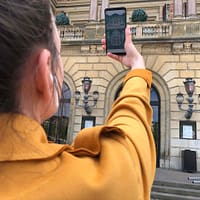 A woman in a yelow coat is atnding in front of the Danish Royal theatre. She is holding up her smartphone