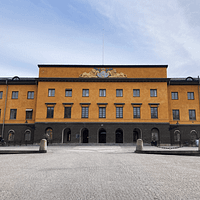 A photograph of Vitterhetsakademiens library in Stockholm. It is a three-story stone building painted a dark ockra color. In front is a large paved plaza.