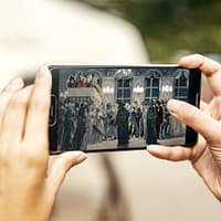 Hands are holding a smartphone. On the screen a scene from the murder of King Gustav the third is visible.