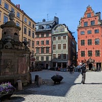 The main square of Stockholm old town. An old well is visible on the left, the surrounding buildings are colorful and the square is paved with cobblestones.