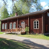 An old, red wooden one-storey school building. It has white trimmed windows and a gravelled lot in front. The are beech trees surrounding it.