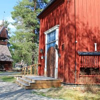 A red church, with a small rectory in the background. the sun is shining and there are trees surrounding the buildings.