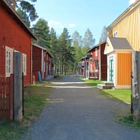A gravelled road is running down between two rows of old wooden buildings. The houses are red and yellow. There is a red picket fence in front.