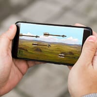 Hands holding a mobile phone. On the screen is an animated image of arrows flying across a battlefield