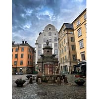 A photo of the old well at Stortorget in Stoclholm Old town. The backdrop is of colorful houses and a gray sky.