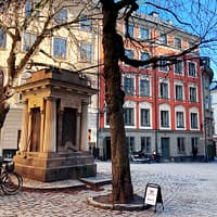 A photo of a little square in Stockholm's old town. In the foreground there is an old well, and in the background are a yellow and a red ston building against a clear blue sky