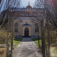 A photograph of the entrance to the German church cemetary. The yellow stone church building is surrounded by a black cast-iron fence with gold details.