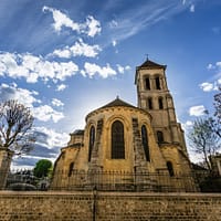 An exterior photo of an old stone church in Paris, with a wall below it and a clear blue sky above.
