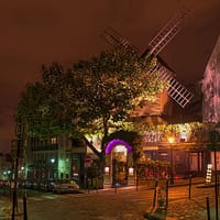 A photo of one of the Montmartre windmills at night. The quiet street is dark, but a spotlight illuminates the small wooden windmill.