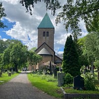A photo of Old Aker church in Oslo. The sandstone church is in the middle of the lush, green cemetery.