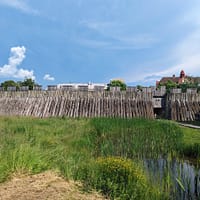Ett foto av Trelleborgen på avstånd. en mängd träpålar utgör borgens palissad och framför syns en gräsyta och en liten bro som går över en damm.