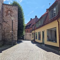 A photo of a Visby street. An old stone wall is visible to the right, and a low, yellow mortared building to the left of a narrow cobblestone street.