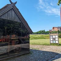 Ett foto av vikingamuseet Trelleborgen. På bilden syns museets kortsida med glasfasad. Innanför glaset skymtas museets utställning, utanför finns ett grönområde och en blå sommarhimmel.