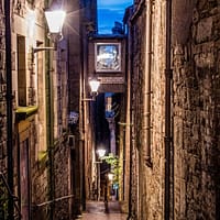 A narrow alley is lit up by a lantern attached to the wall. Steep steps descend into darkness