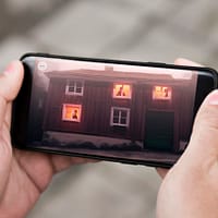 A close up photo of hand holding a smartphone. on the screen is an illustration of a haunted house with ghosts in the windows.