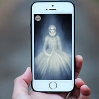 A close up of a hand holding a smartphone. On the screen is an illustration of a ghost queen.