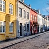 A photo of one of the street in the Nyboder area of Copenhagen. Low. colorful stone house line a narrow cobble stone street with plants and trees along the sidewalk.