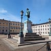 A photograph of the Gustaf Adolf square in Gothenburg. A bronze statue of Gustaf Adolf, pointing down at the square. The surrounding buildings are elegant stone buildings in muted colors.