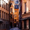 A photo of a Stockholm Old town alley. Sun is gleaming down on mortared buildings painted shades of yellow and orange. A cobblestone street runs between them.