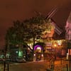 A photo of one of the Montmartre windmills at night. The quiet street is dark, but a spotlight illuminates the small wooden windmill.