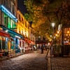 A photo of the Place du Tertre in Paris at night. Small bistros are lining both sides of a cozy square. There are ambient light and seating outside on the curb.