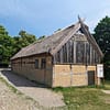 Ett foto av det rekonstruerade medeltida korsvirkeshuset vid Trelleborgens museumsområde. Huset är ljusgrått, ljusgult och avlångt.