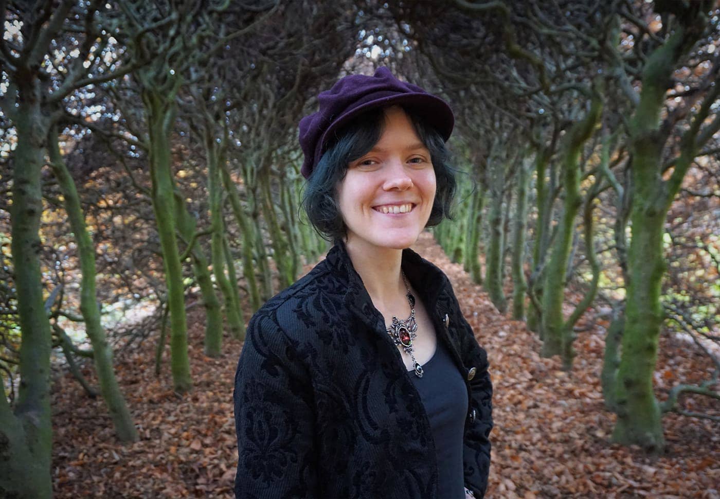 A portrait image of Marie Metso, who is standing in a clearing in the woods. She is wearing a beret and smiling at the camera.