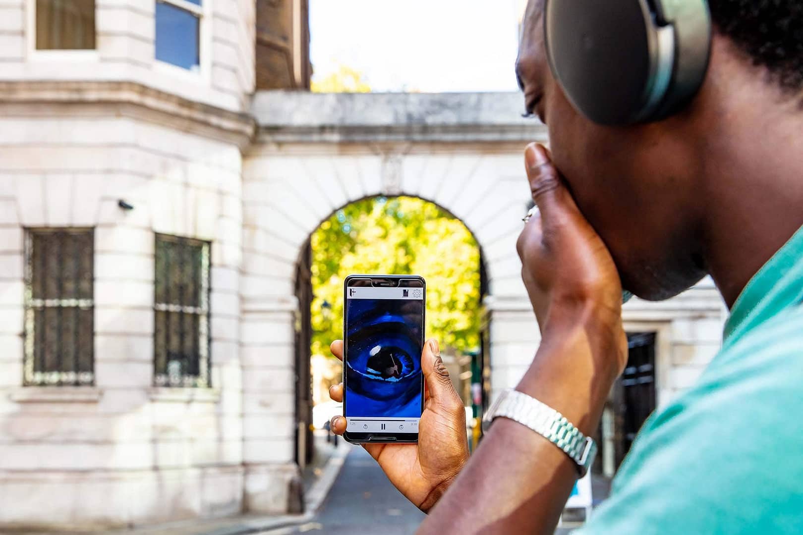 A young man is standing in front of a white stone building in London. He is holding a smartphone, looking at the screen with an amazed expression