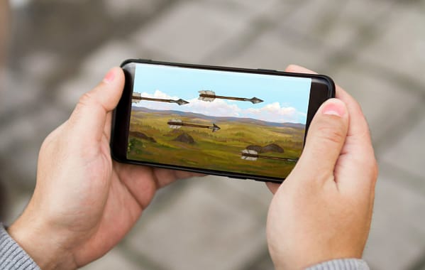 Hands holding a mobile phone. On the screen is an animated image of arrows flying across a battlefield