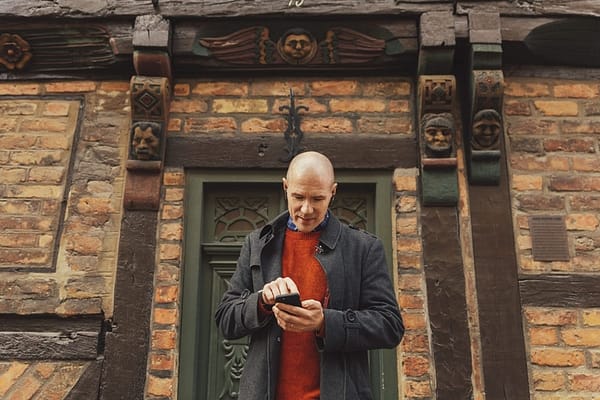 A man is standing in front of a half-timbred house. He is looking at his phone