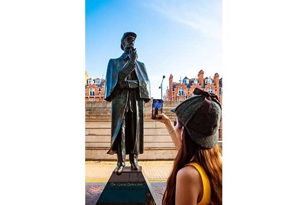 A young woman in a deerstalker hat is taking a picture of a Sherlock Holmes statue