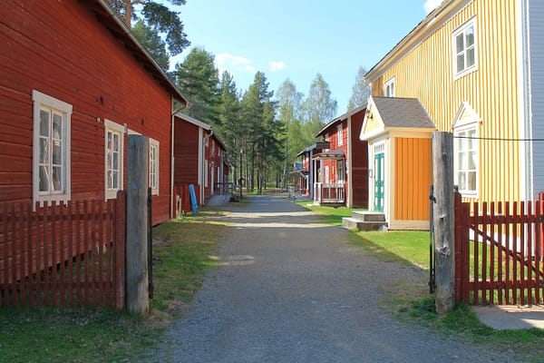 A gravelled road is running down between two rows of old wooden buildings. The houses are red and yellow. There is a red picket fence in front.