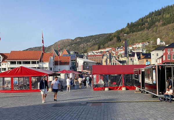 A photo of the Fisketorget square in Bergen, Norway. There are red little stalls around the square and people are walking in the sunshine.