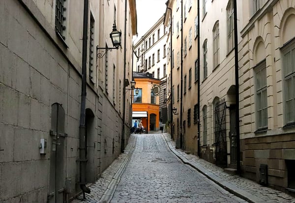 A photo of a Stockholm Old town alley. The alley is dark and dreary, with the sun and a colorful building visible at the very end of it.