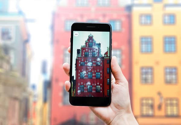 A close up photo of hands holding a smartphone. On the screen is an image of a haunted building, with skulls floating around in the air.