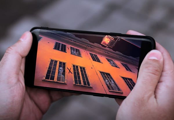 A close up photo of hands holding a smartphone. On the screen is a picture of a haunted building, with ghosts in the windows.