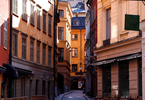 A photo of a Stockholm Old town alley. Sun is gleaming down on mortared buildings painted shades of yellow and orange. A cobblestone street runs between them.