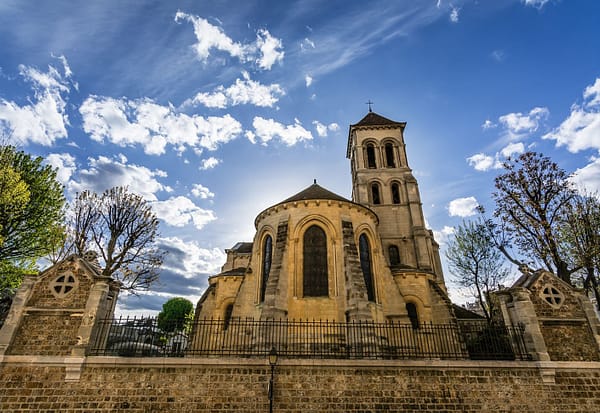 An exterior photo of an old stone church in Paris, with a wall below it and a clear blue sky above.