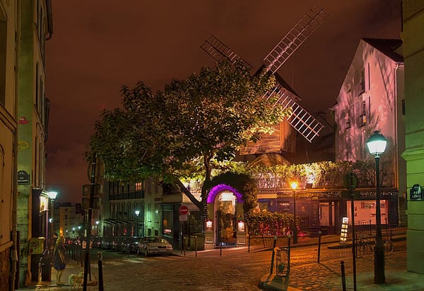 A photo of one of the Montmartre windmills at night. The quiet street is dark, but a spotlight illuminates the small wooden windmill.