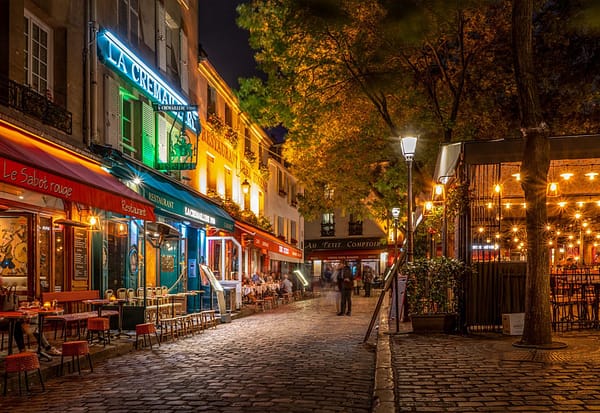 A photo of the Place du Tertre in Paris at night. Small bistros are lining both sides of a cozy square. There are ambient light and seating outside on the curb.