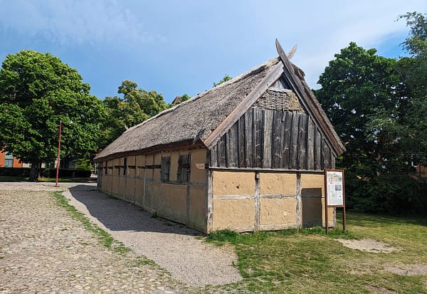 Ett foto av det rekonstruerade medeltida korsvirkeshuset vid Trelleborgens museumsområde. Huset är ljusgrått, ljusgult och avlångt.