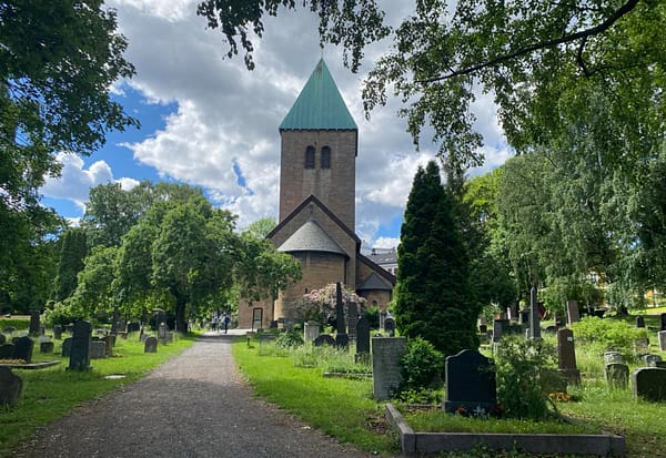 A photo of Old Aker church in Oslo. The sandstone church is in the middle of the lush, green cemetery.