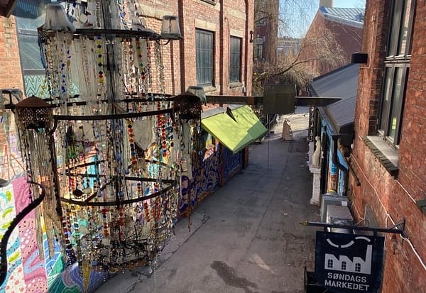 A photo taken from above Ingensteds alley in Oslo. Old factory buildings covered in graffiti have been turned into a market area. A glass chandeleir is hanging in the middle of the alley.