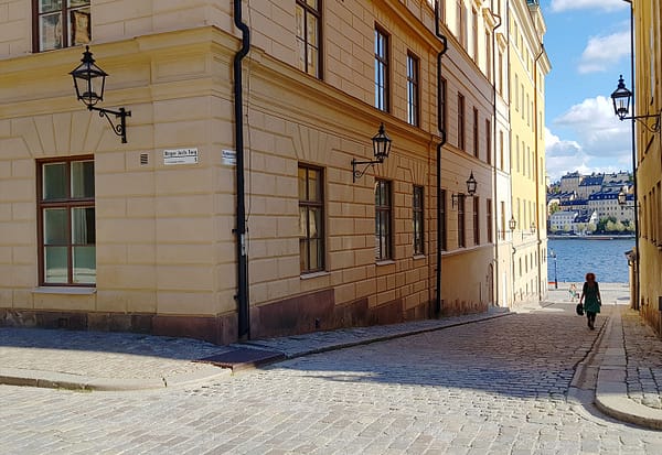 A photo of a street on Riddarholmen in Stockholm. Yellow, mortared buildings are lining a narrow cobblestone street. At the end of the street you can see clear blue sky and water.