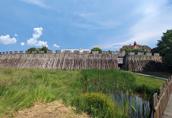 Ett foto av Trelleborgen på avstånd. en mängd träpålar utgör borgens palissad och framför syns en gräsyta och en liten bro som går över en damm.