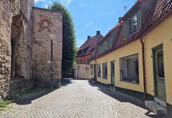 A photo of a Visby street. An old stone wall is visible to the right, and a low, yellow mortared building to the left of a narrow cobblestone street.