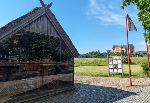 Ett foto av vikingamuseet Trelleborgen. På bilden syns museets kortsida med glasfasad. Innanför glaset skymtas museets utställning, utanför finns ett grönområde och en blå sommarhimmel.