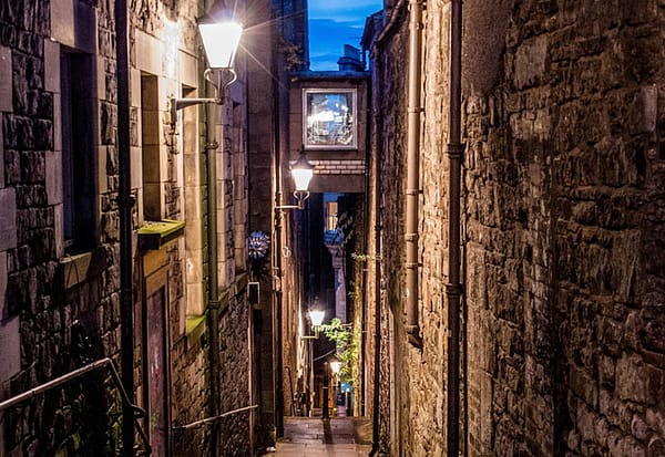 A narrow alley is lit up by a lantern attached to the wall. Steep steps descend into darkness